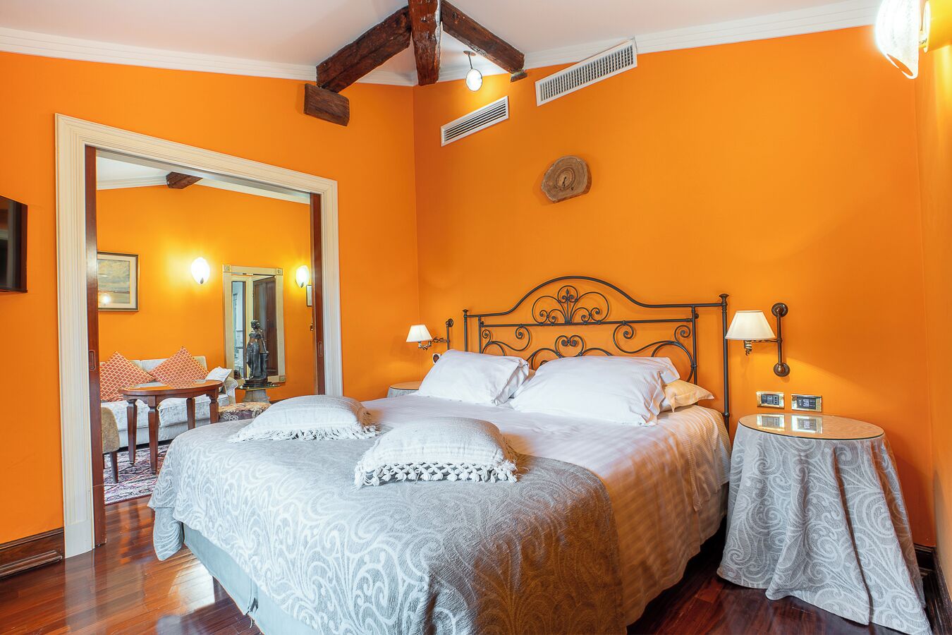 A bedroom with a large bed, a wooden headboard, two bedside tables with lamps, a mirror, and a wooden floor.