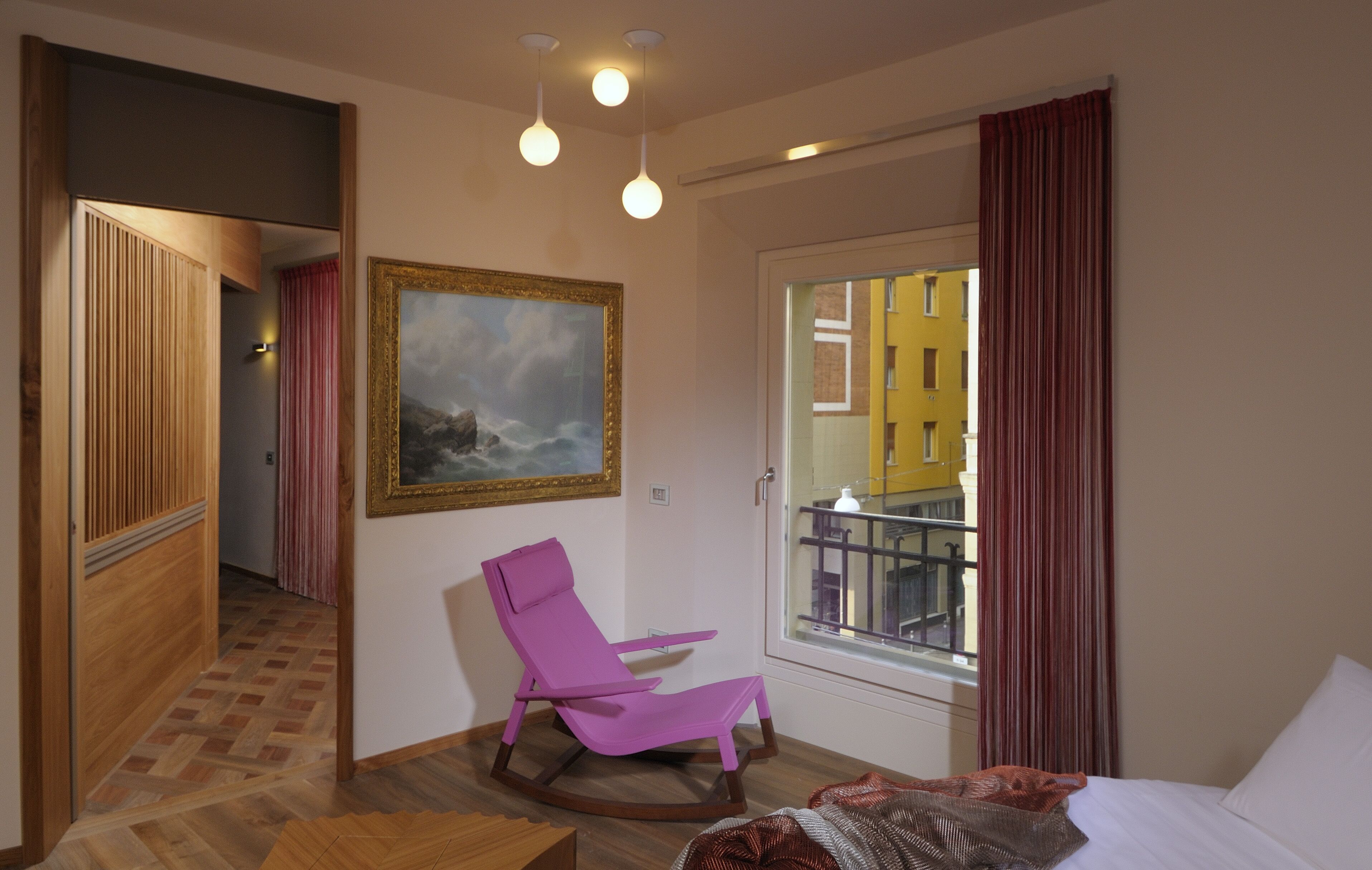 A hotel room with a wooden floor, a pink chair, a painting, a window with curtains, and a bed.