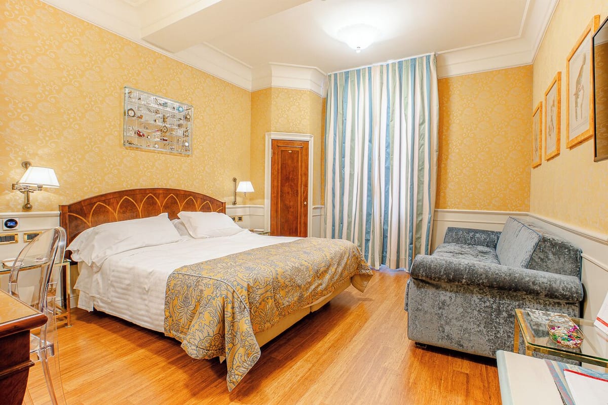 Une chambre d’hôtel comprenant un lit, une chaise, un bureau, un miroir, une fenêtre avec des rideaux et un mur orné de tableaux.