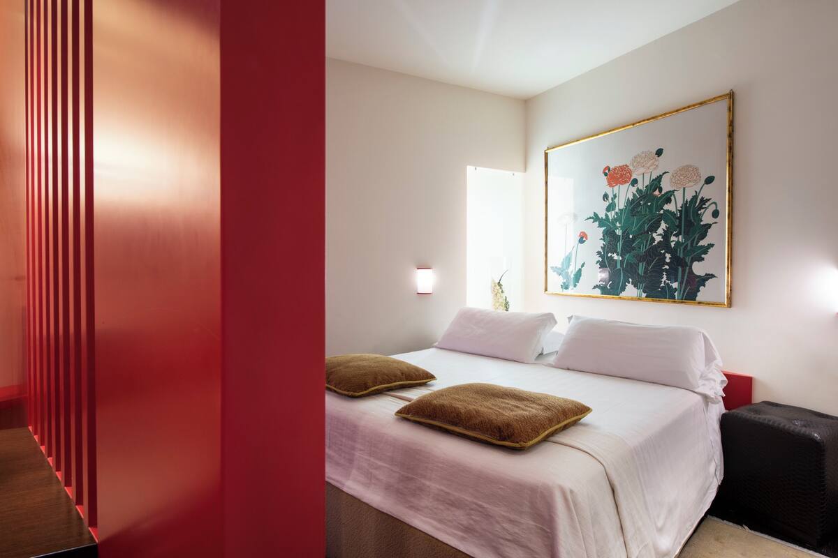 Une chambre d’hôtel avec un lit, des oreillers, un tableau, une table de chevet et un mur rouge.