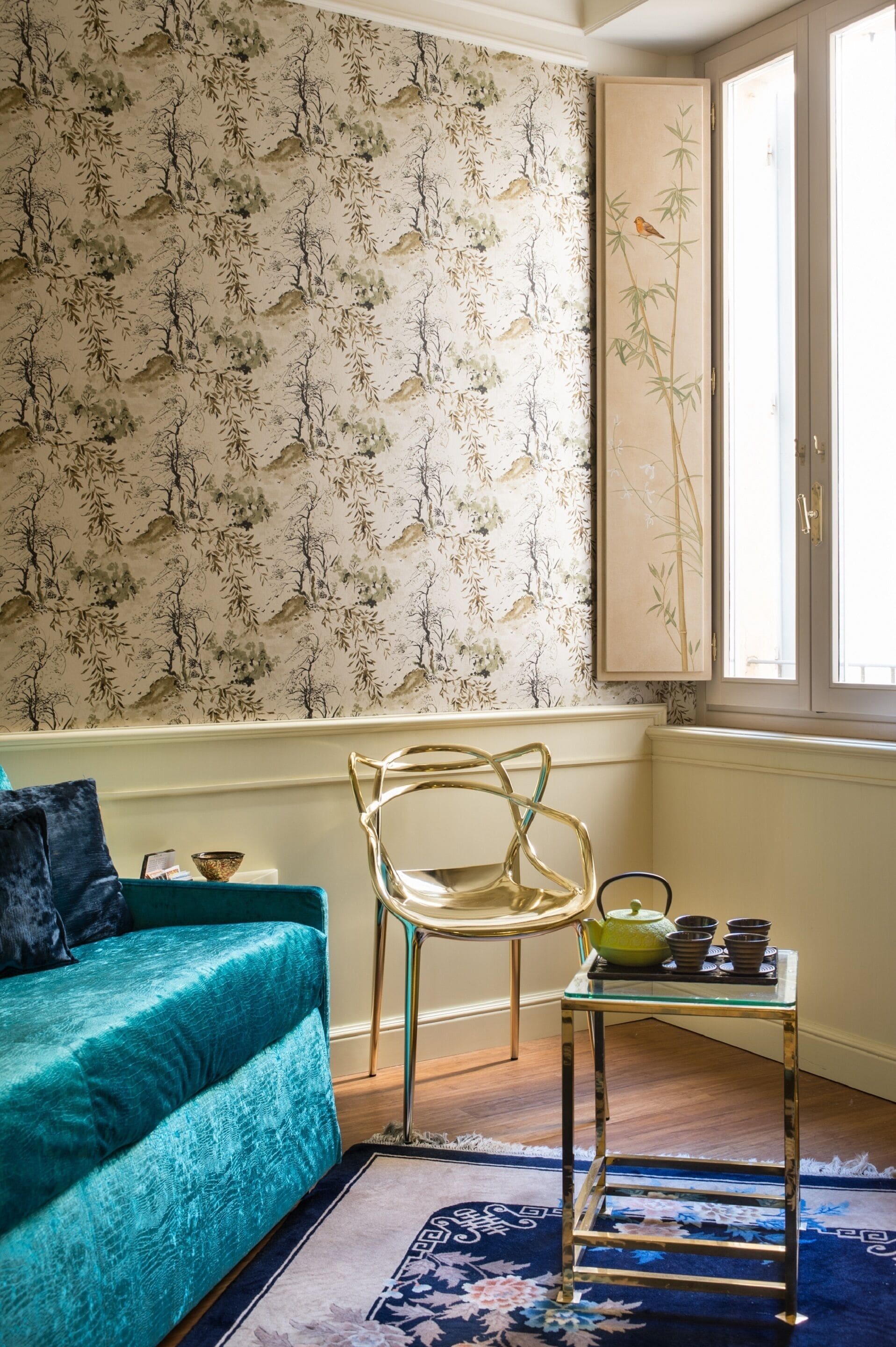A room with a teal sofa, a glass table with a green teapot, and a floral-patterned wallpaper.