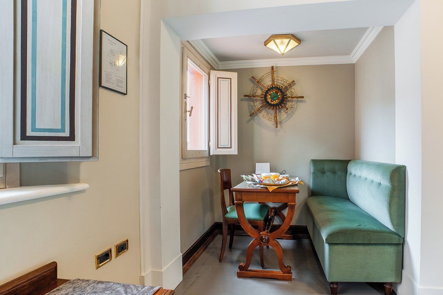Un pasillo estrecho con un sofá verde, una silla de madera, una mesa con papeles y una taza, un reloj de pared y un cuadro enmarcado.