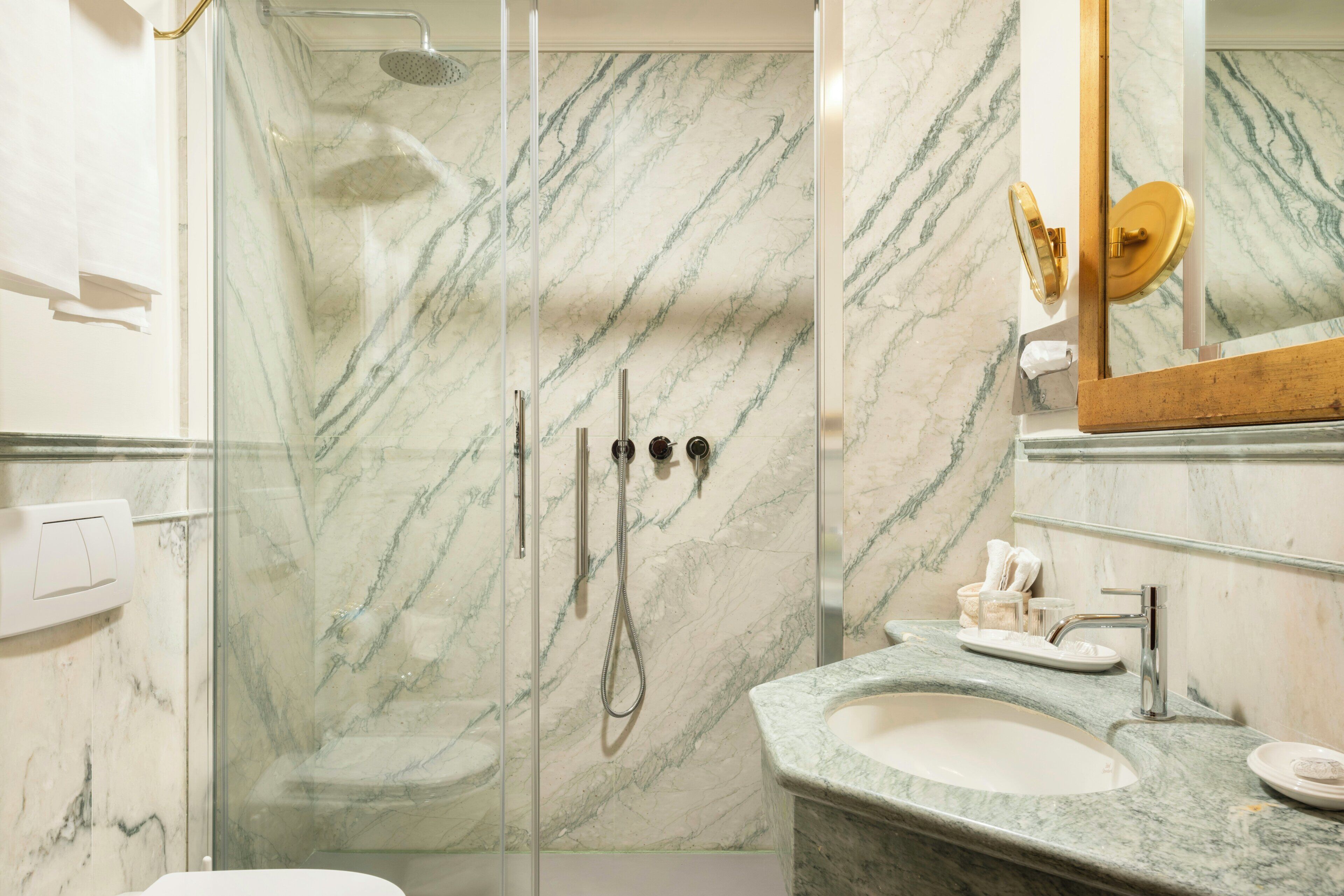 A bathroom with a glass shower enclosure, a sink with a marble countertop, and a mirror with a gold frame.