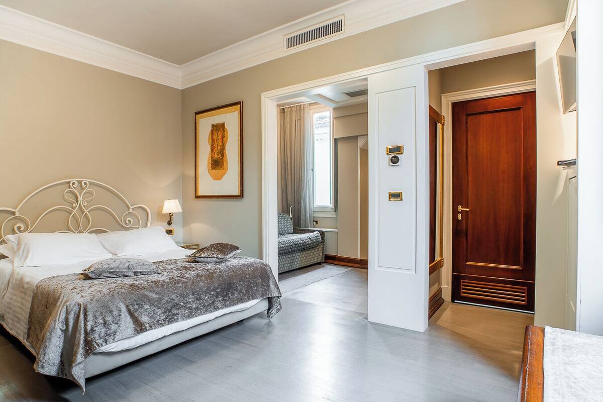 Une chambre d’hôtel avec un lit, une table de chevet, une lampe, un tableau encadré au mur et une porte donnant sur une autre pièce.