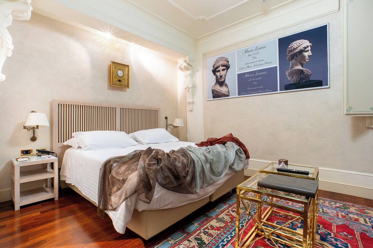 Une chambre d’hôtel avec un lit, une table de chevet, une lampe, une horloge, des affiches et une petite table avec un tapis.