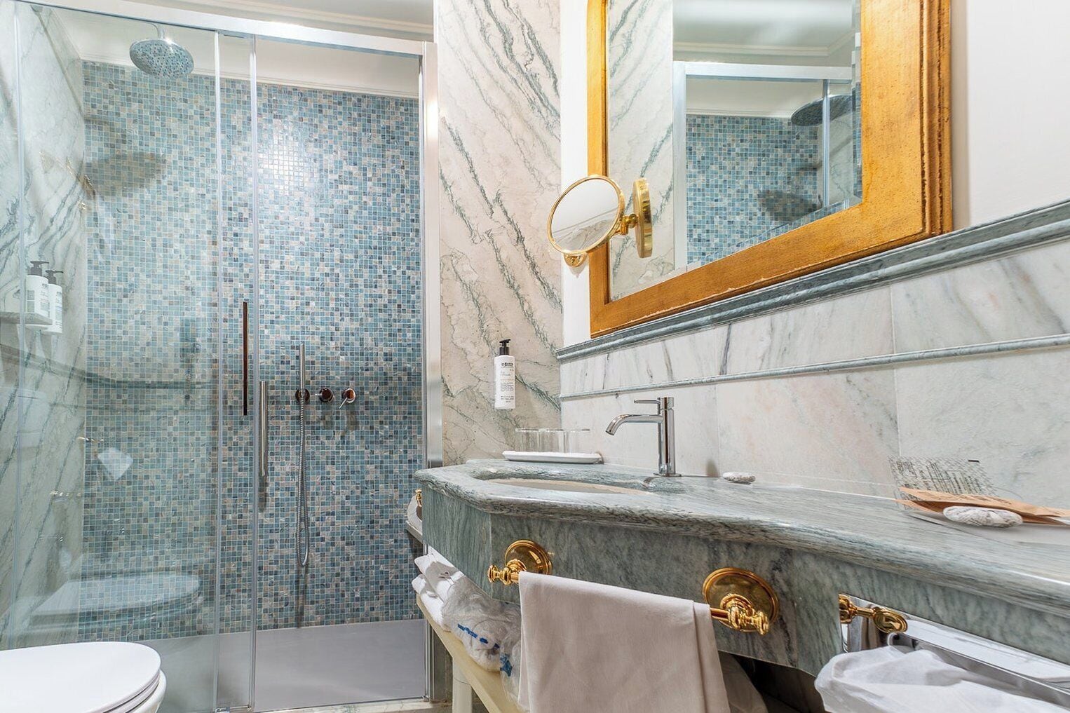 Un baño con ducha envuelta en vidrio, un lavamanos de mármol y un espejo con marco dorado.