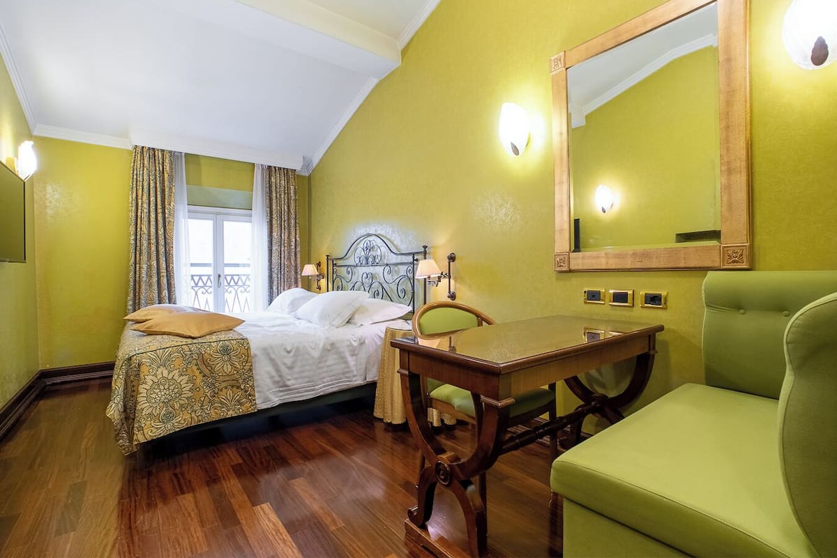 Une chambre d’hôtel avec un lit, une table, une chaise, un miroir et une fenêtre avec des rideaux.