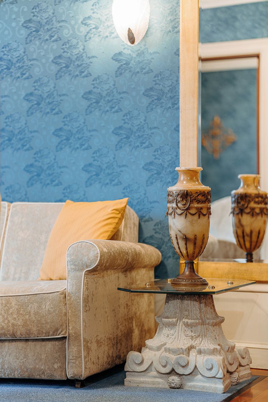 Un sofá antiguo con un cojín, un jarrón decorativo sobre una mesa de vidrio y un espejo que refleja la habitación.