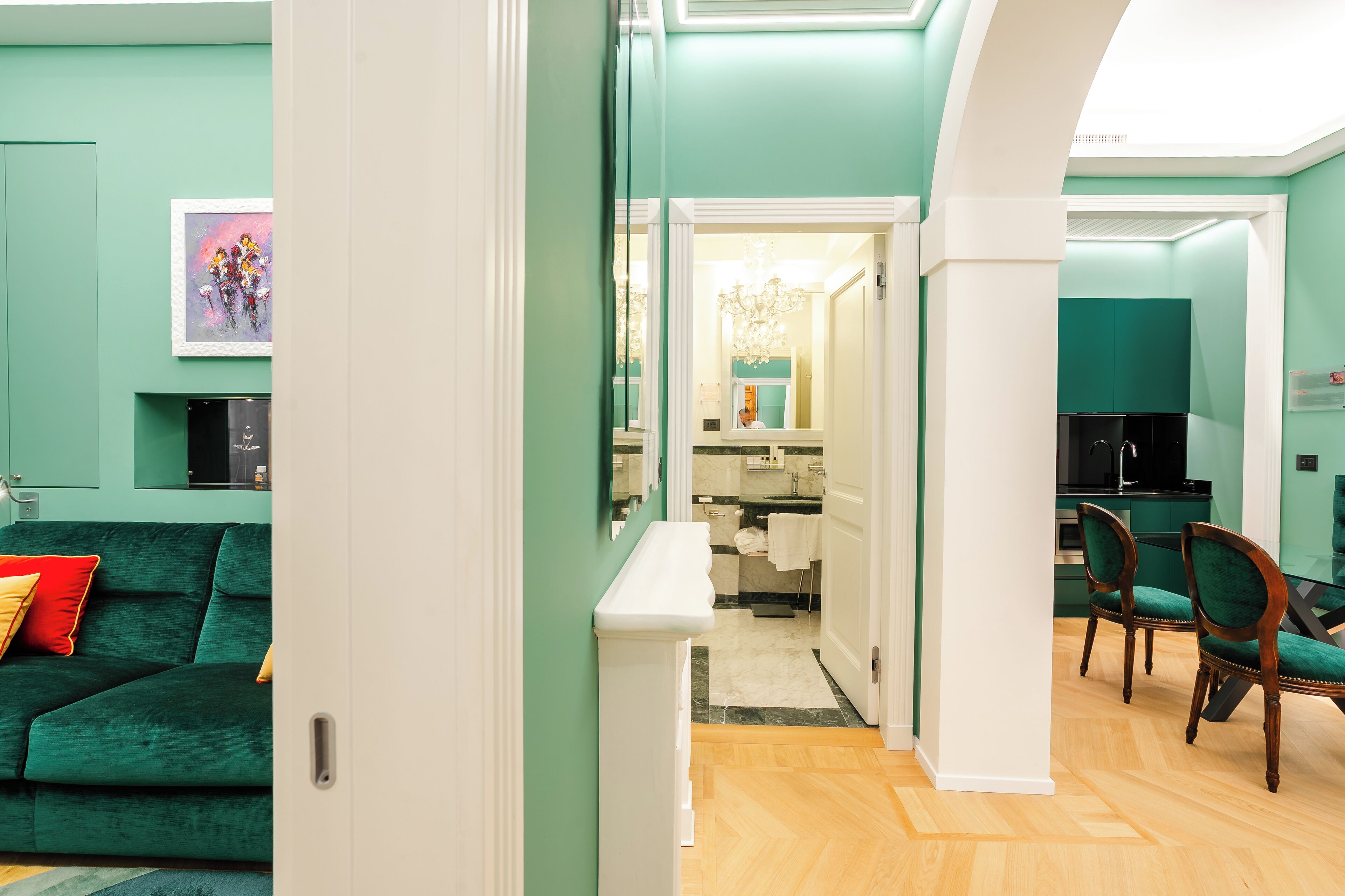A modern living room with a green sofa, a dining area with two chairs, and a kitchen visible through an open door.