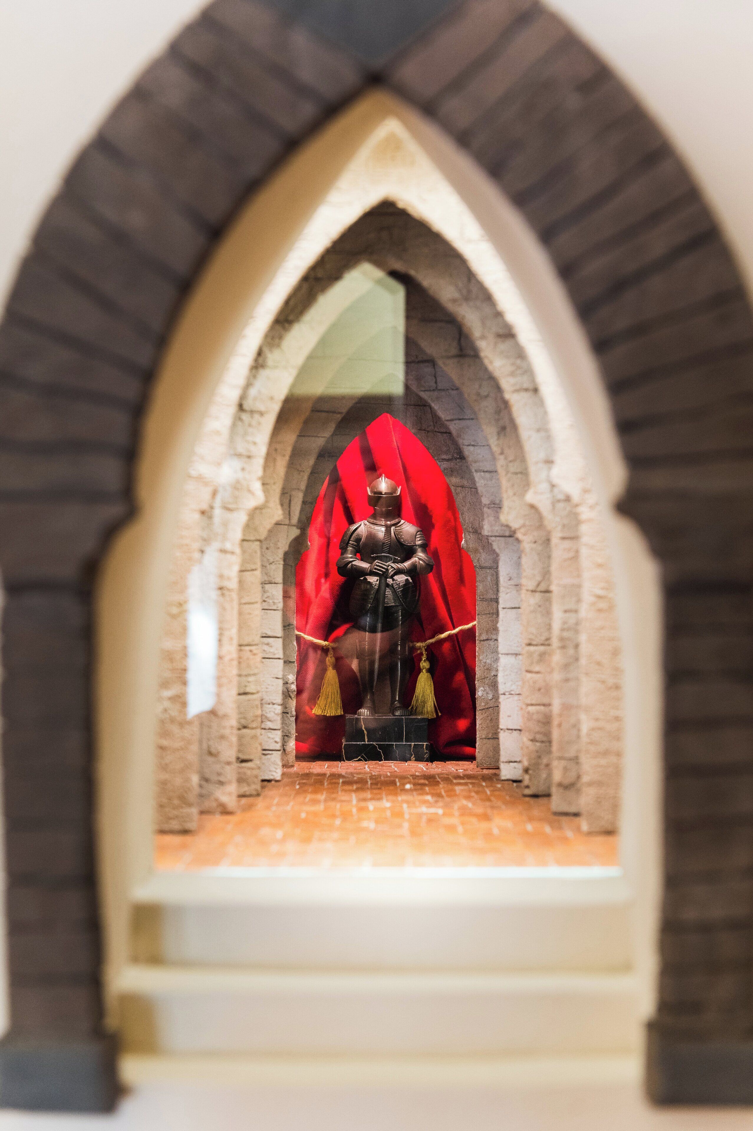 Un modello di cavaliere in armatura seduto su un trono, circondato da un drappo rosso, all'interno di un arco in pietra.