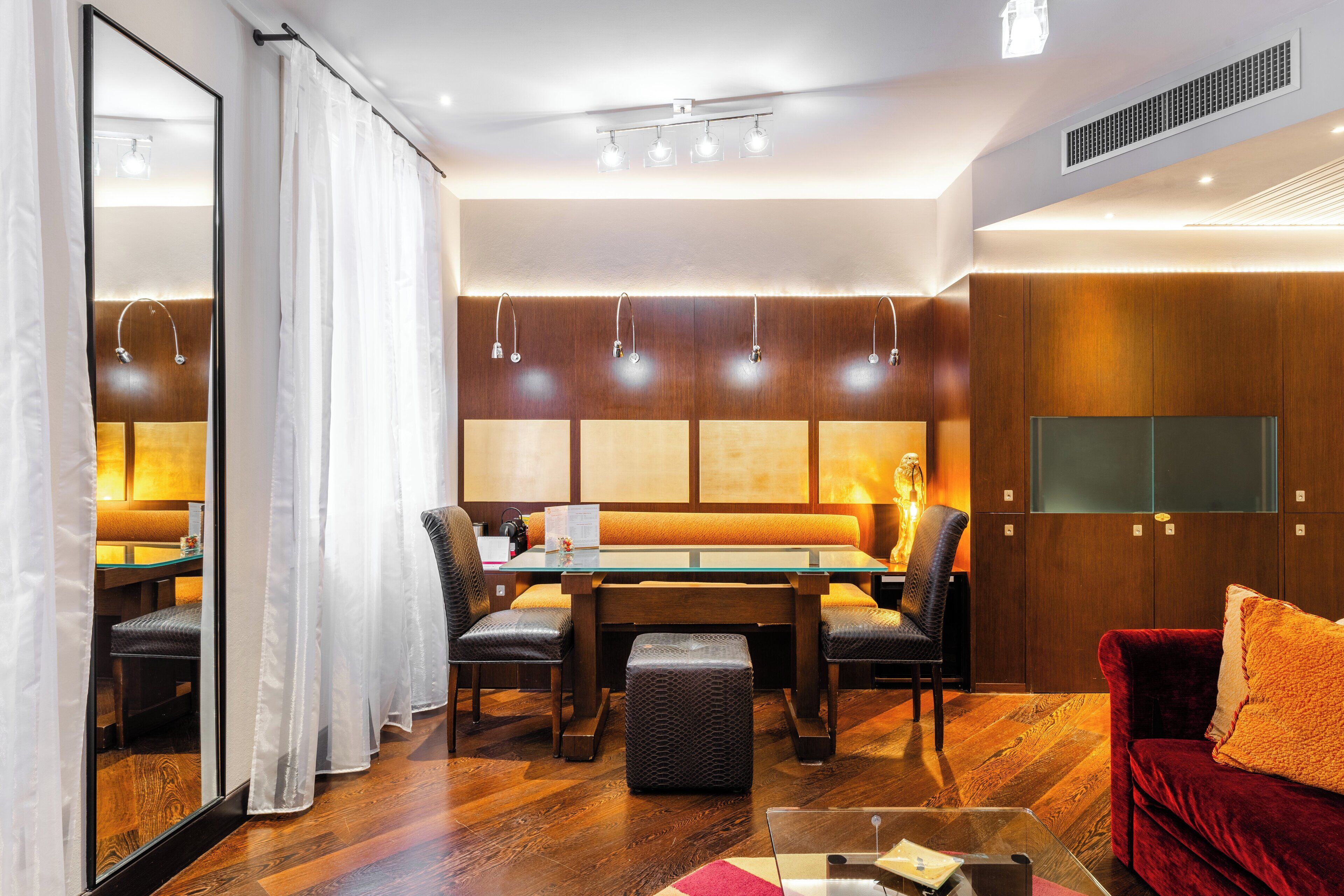 A modern hotel room with a wooden floor, a glass coffee table, a red sofa, a dining table with chairs, and a large mirror.