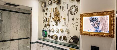 A bathroom with a marble shower, a glass-topped vanity, and a wall display of clocks and decorative items.