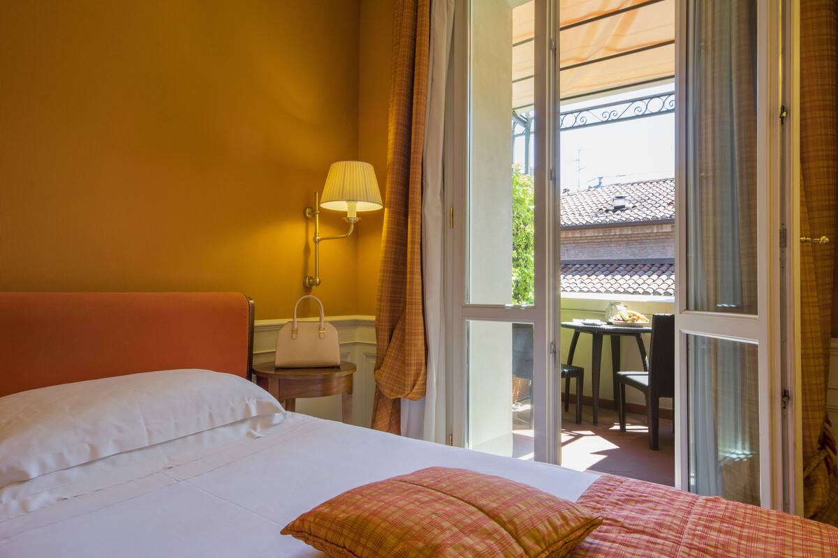 A hotel room with a bed, a nightstand, a lamp, a window with curtains, and a view of the outside balcony.