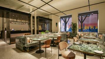 Um restaurante moderno com mesas de mármore e assentos acolchoados, amplas janelas com vista para a cidade e um espaço bar com prateleiras de vidro.