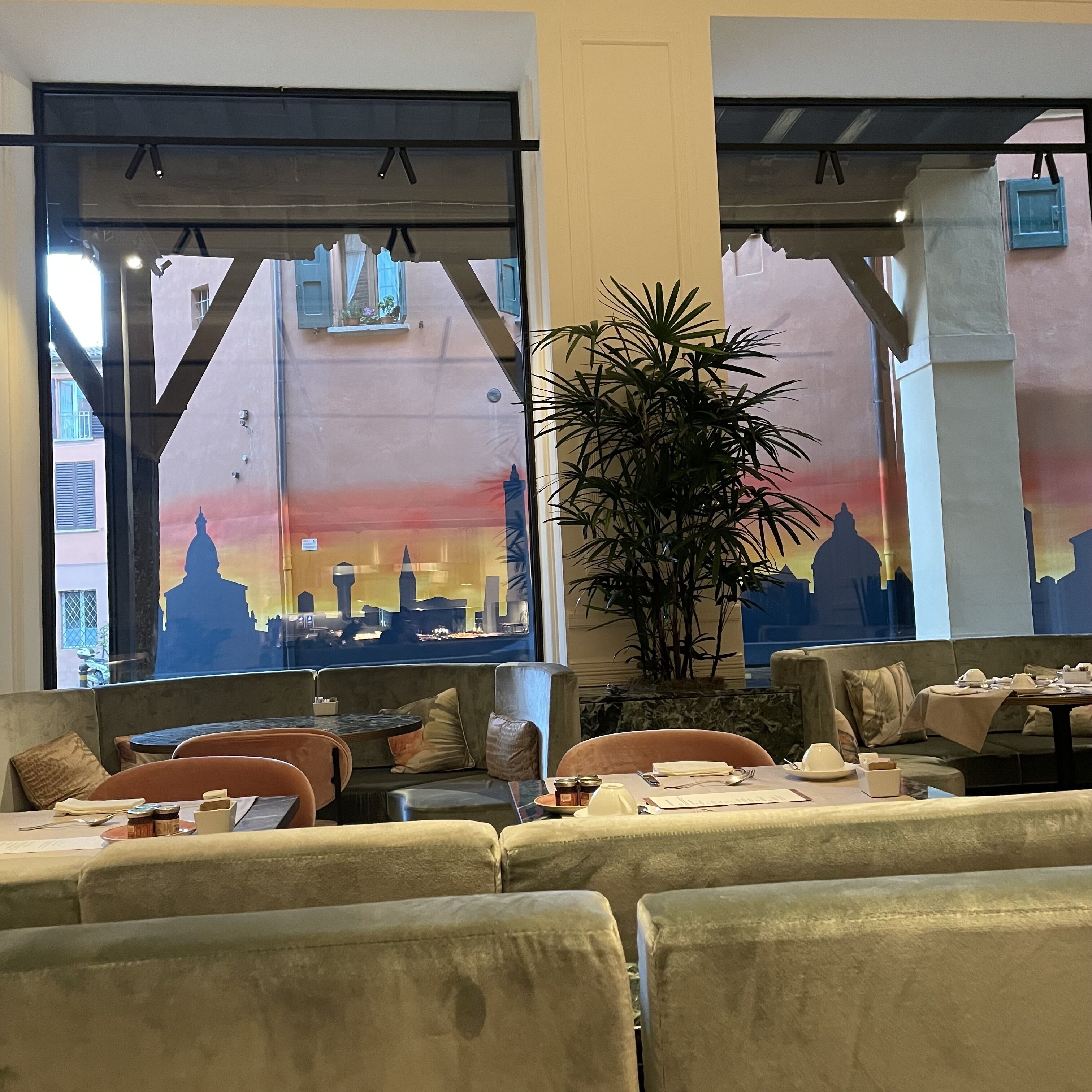 A restaurant interior with a cityscape mural on the wall, cushioned seating, and a large potted plant.