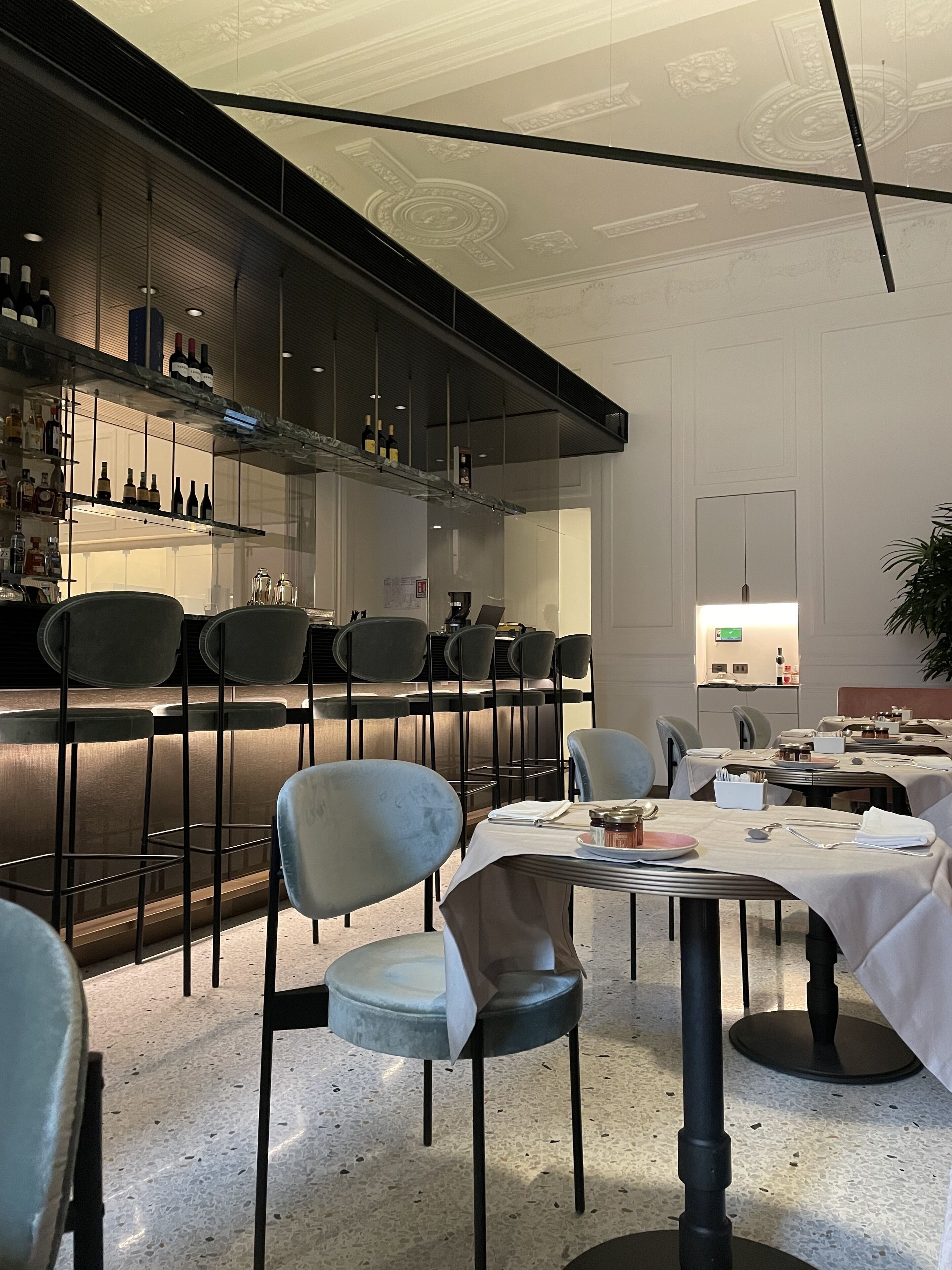 A modern restaurant interior with bar seating, tables set for dining, and a bar area with bottles and glasses.