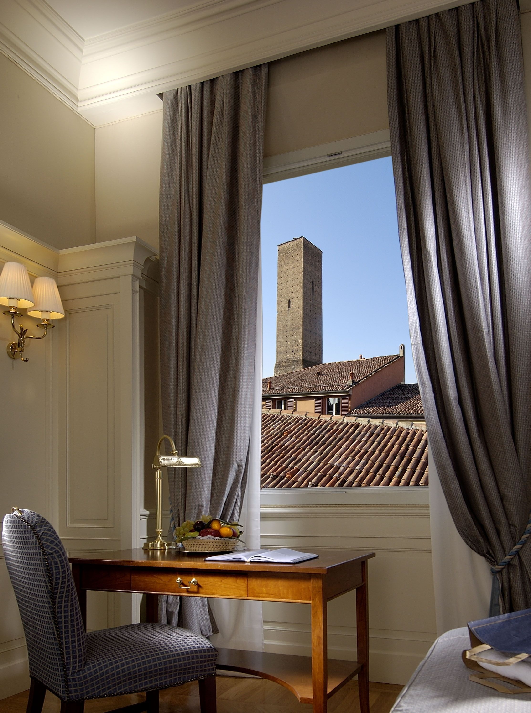 A hotel room with a wooden desk, a chair, a window with curtains, a lamp, and a view of a tower and tiled roof.