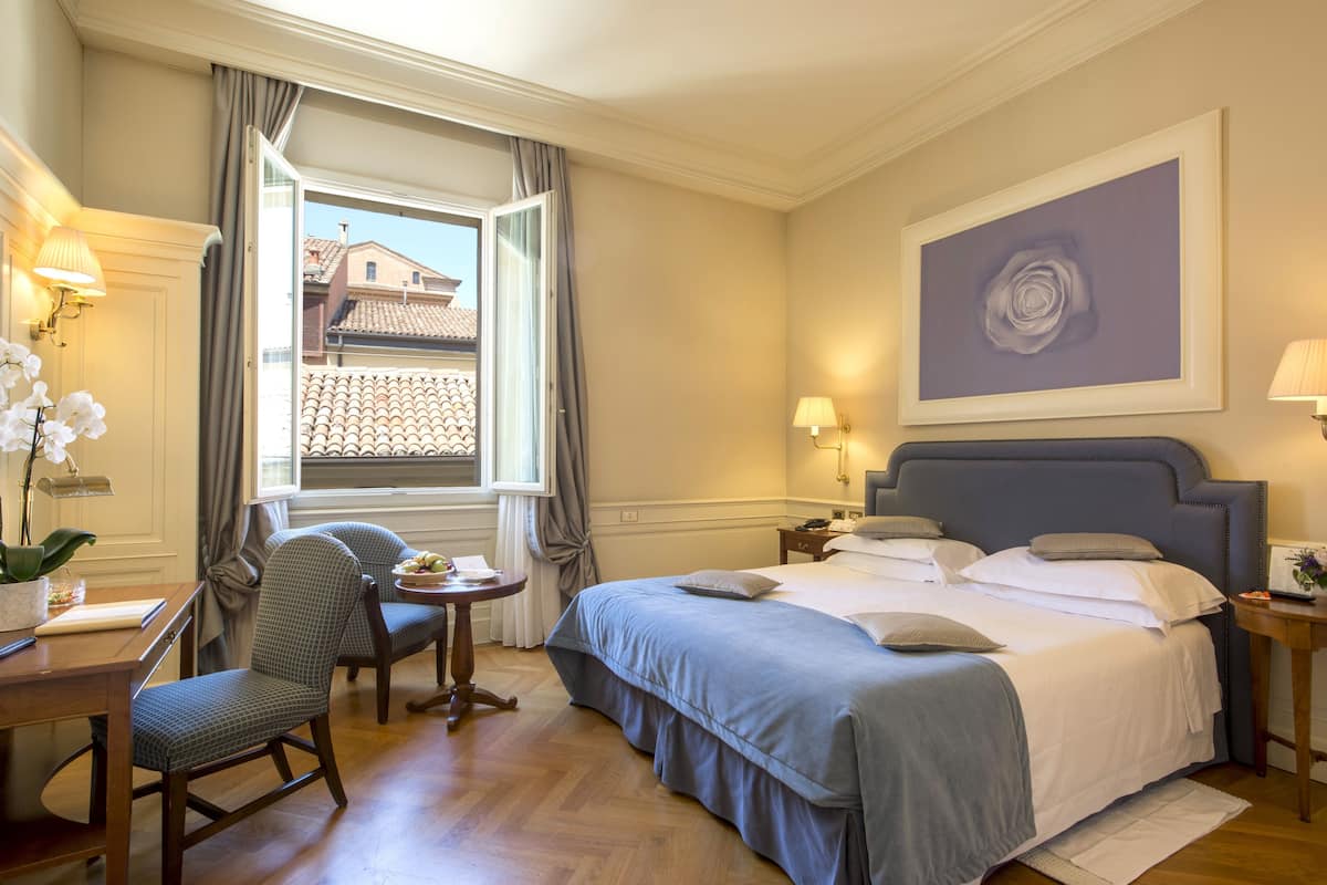 A hotel room with a bed, a desk, a chair, a window with curtains, and a painting on the wall.