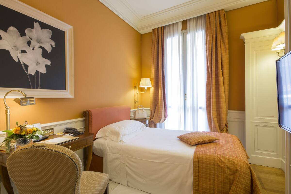 A hotel room with a bed, a desk with a lamp, a chair, a television, and a painting of flowers on the wall.