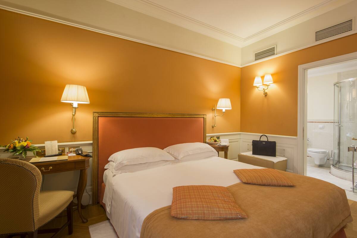 A hotel room with a large bed, two bedside lamps, a desk with a chair, a small table with a handbag, and a bathroom with a glass shower.
