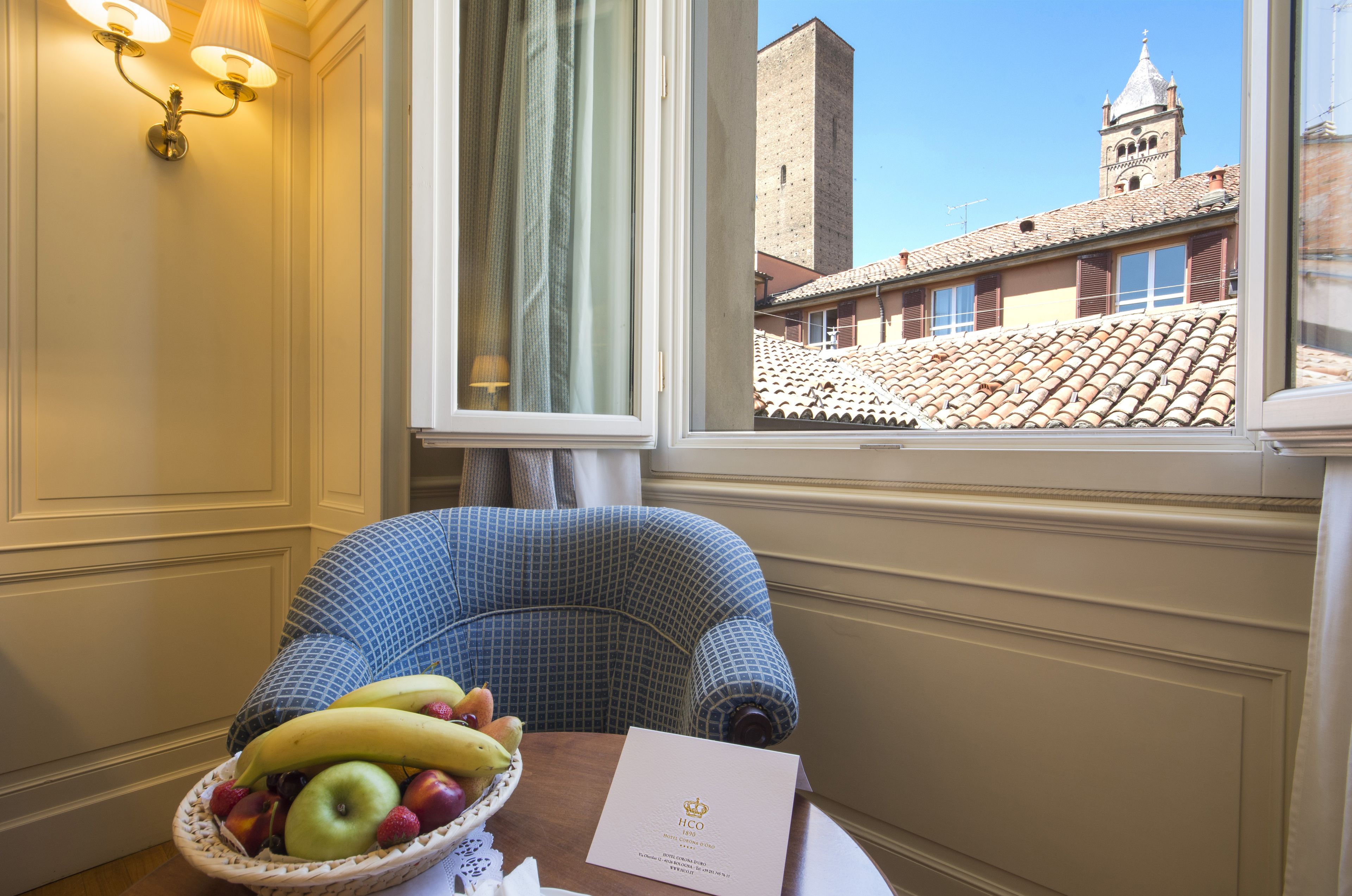 A room with a view of a historic building, a wicker fruit basket on a table, and a blue patterned chair.