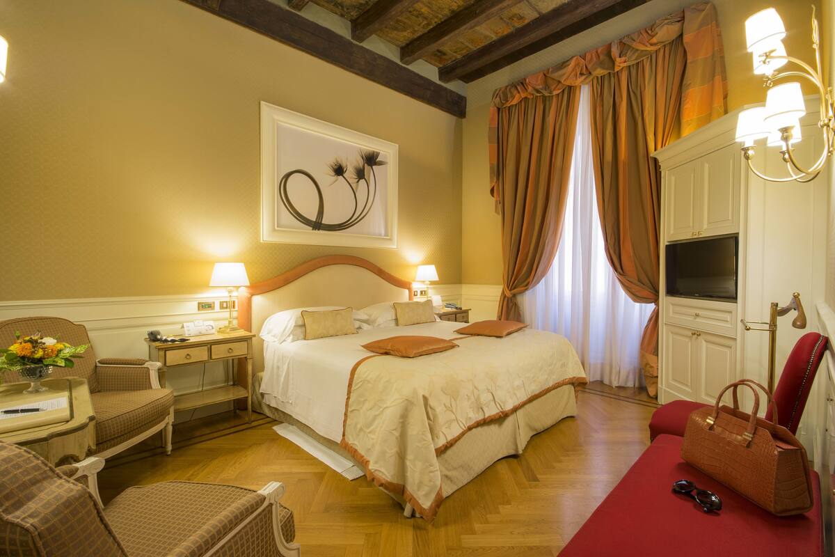 A hotel room with a large bed, two bedside tables with lamps, a chair, a desk, a TV, and a red rug.