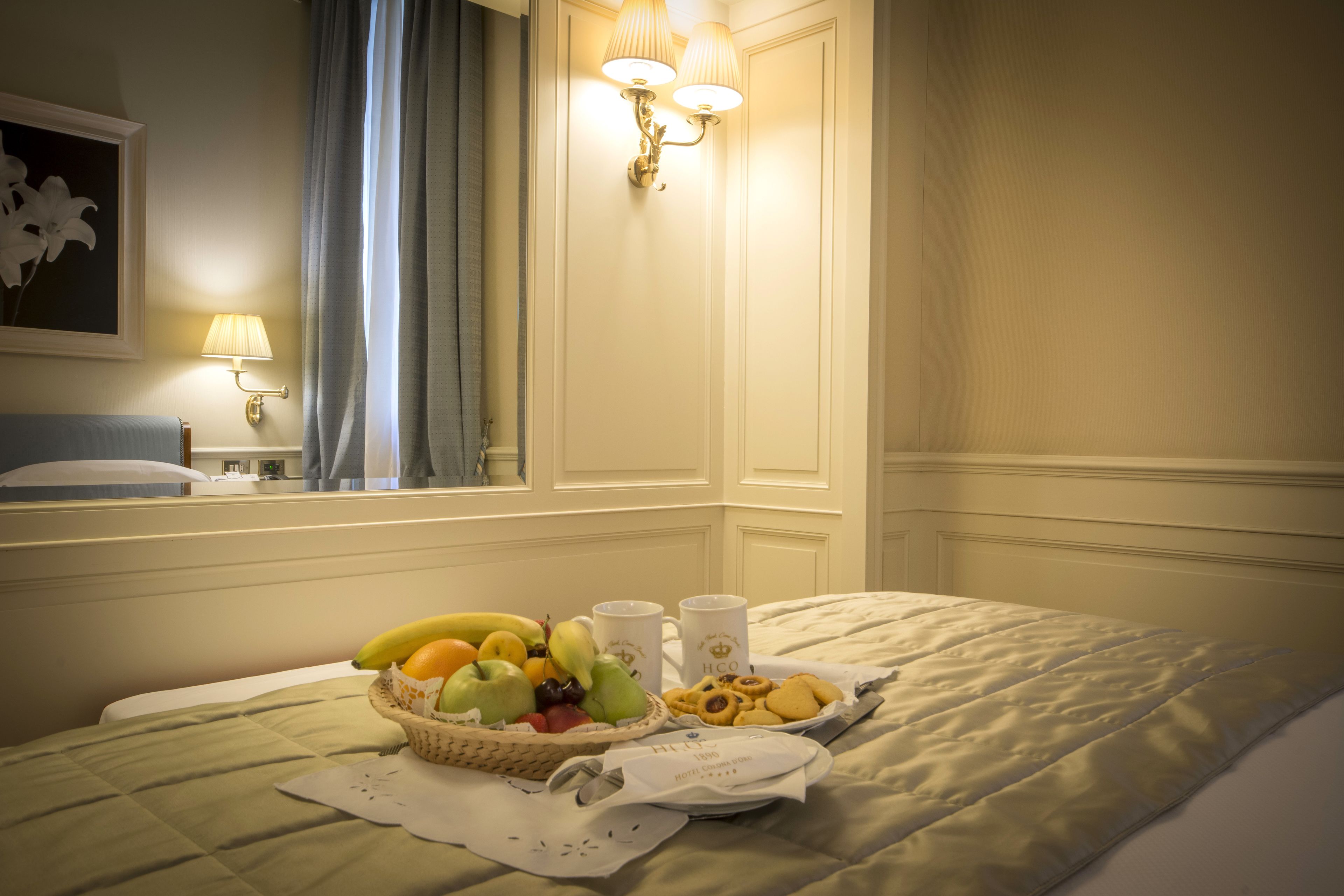 Una cama bien hecha con una cesta de fruta, galletas y dos tazas sobre una mesa junto a ella.