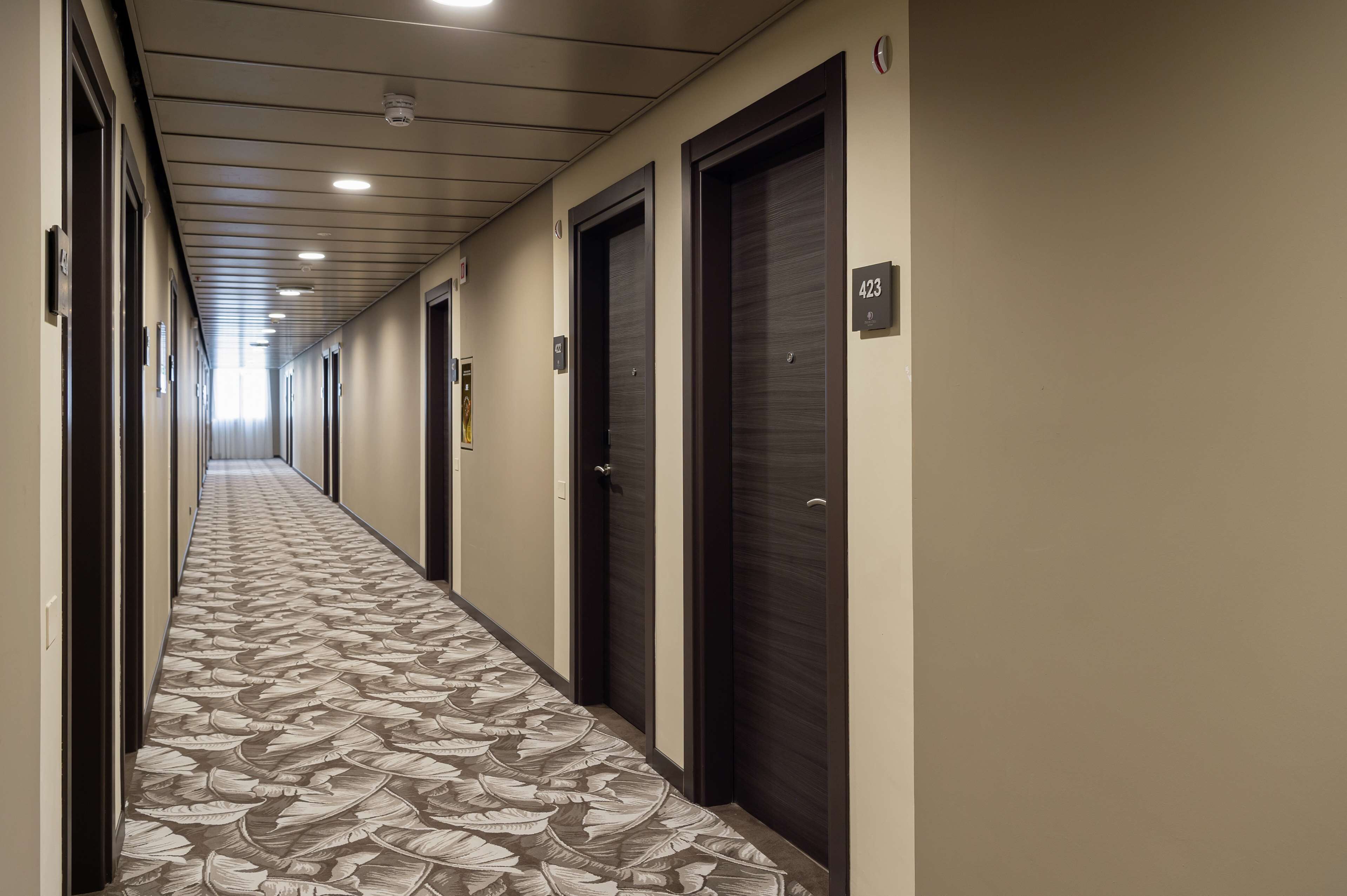 A long, narrow hallway with a patterned carpet, dark doors, and numbered room signs.