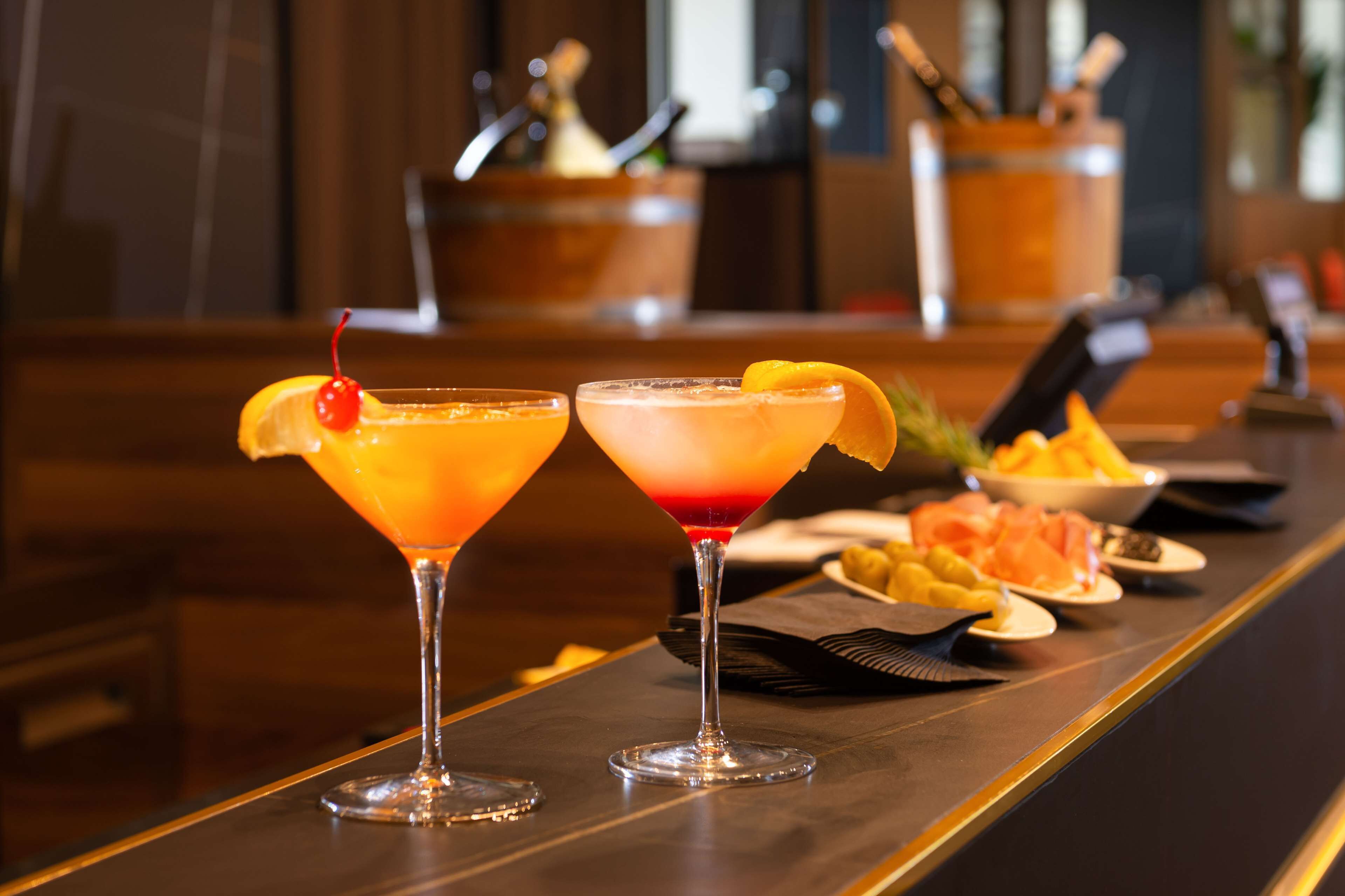 Two cocktails on a bar counter with a wooden background and a blurred bar area in the background.