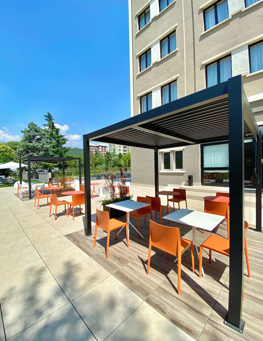 Eine Außenpatio mit Tischen und Stühlen, einer Pergola und einem Gebäude im Hintergrund.