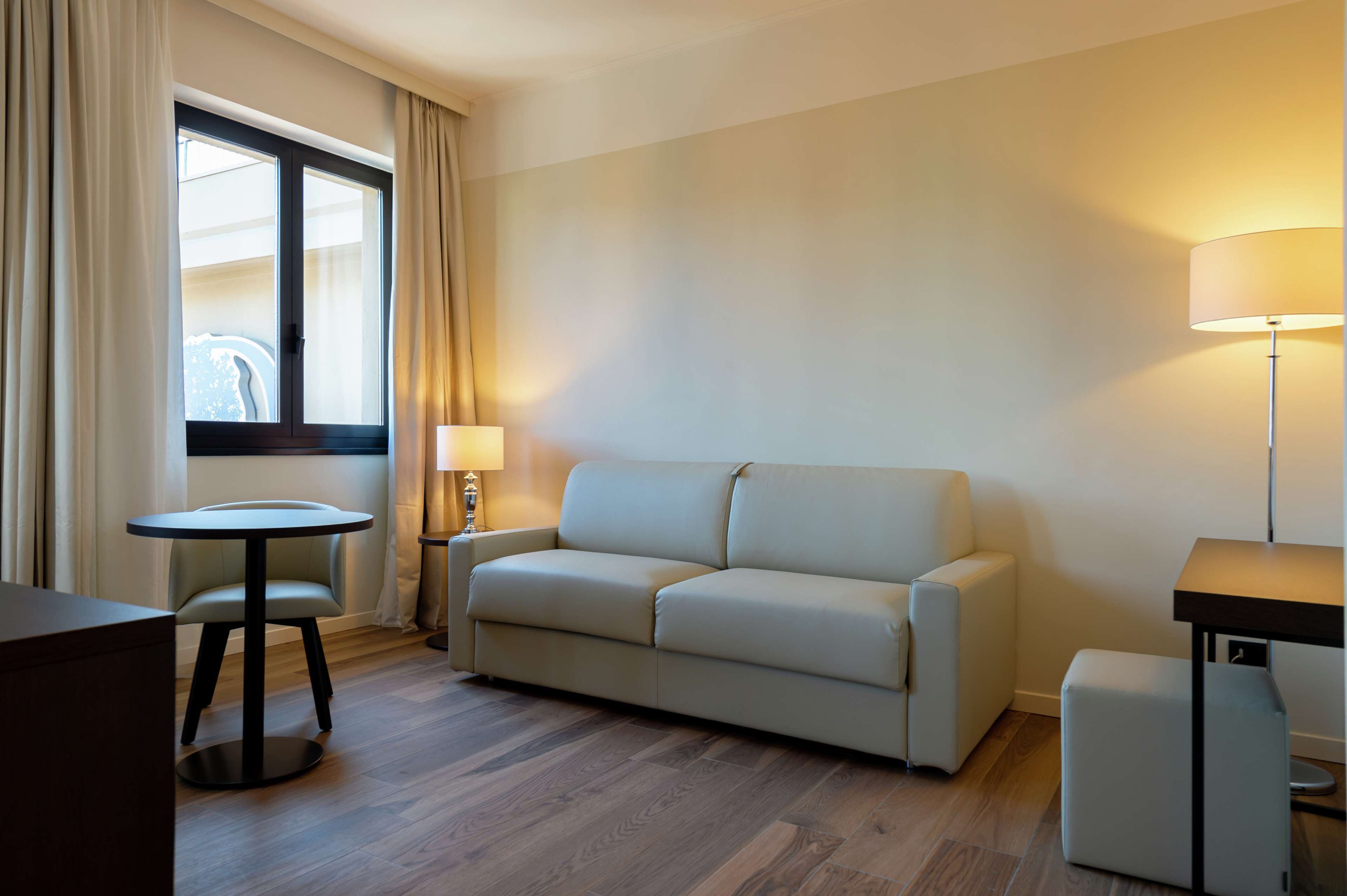 Una camera d'albergo con un divano, un tavolino rotondo, una lampada e una finestra con tende.