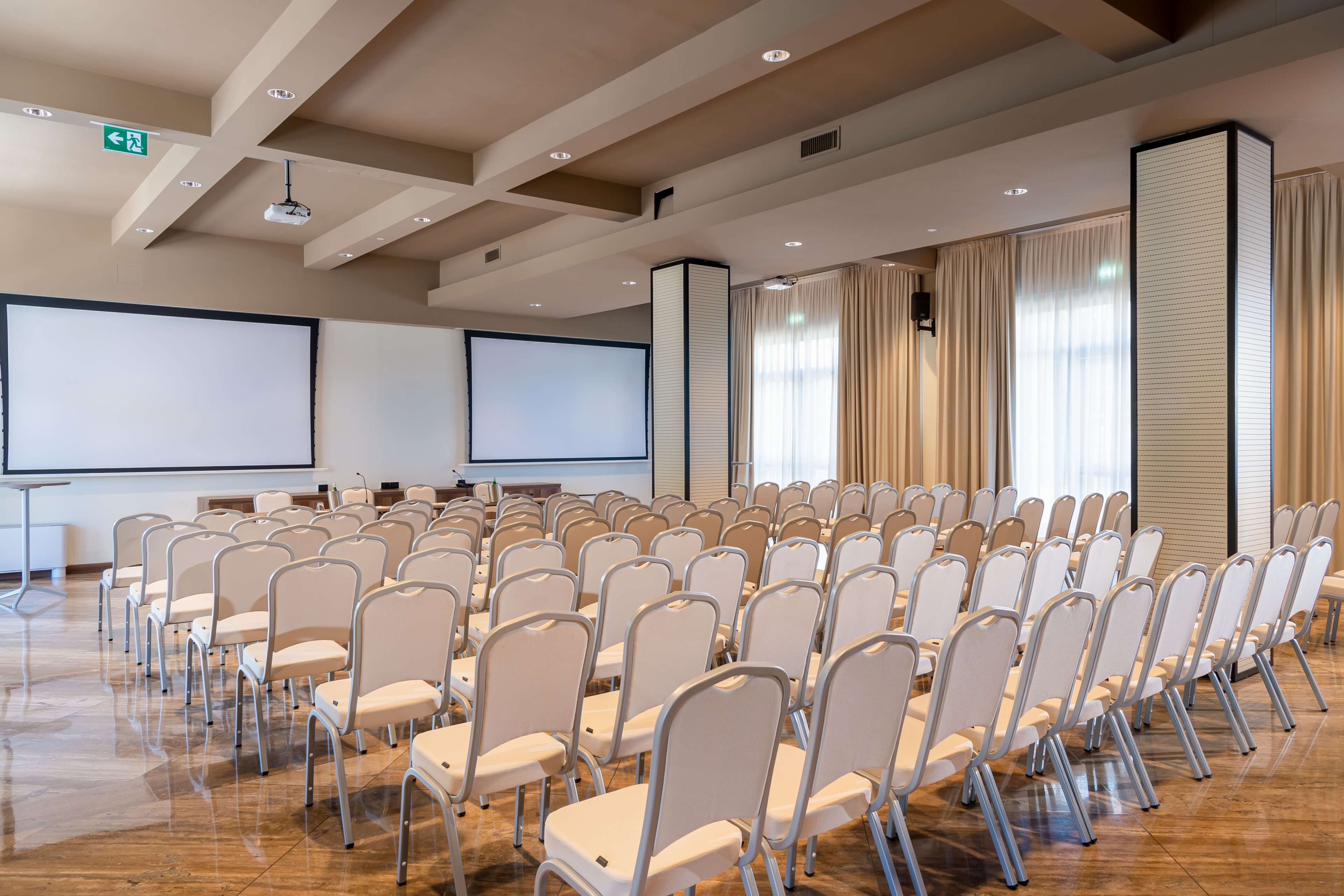 Un salón de conferencias con hileras de sillas blancas, una pantalla grande y un proyector.