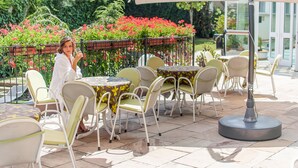 Un patio al aire libre con mesas y sillas redondas, rodeado de macetas con plantas y un jardín.