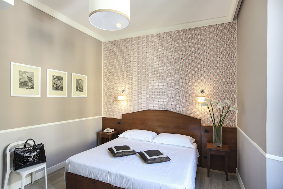A neatly arranged hotel room with a large bed, bedside lamps, a nightstand with a phone, and a small table with a vase of flowers.
