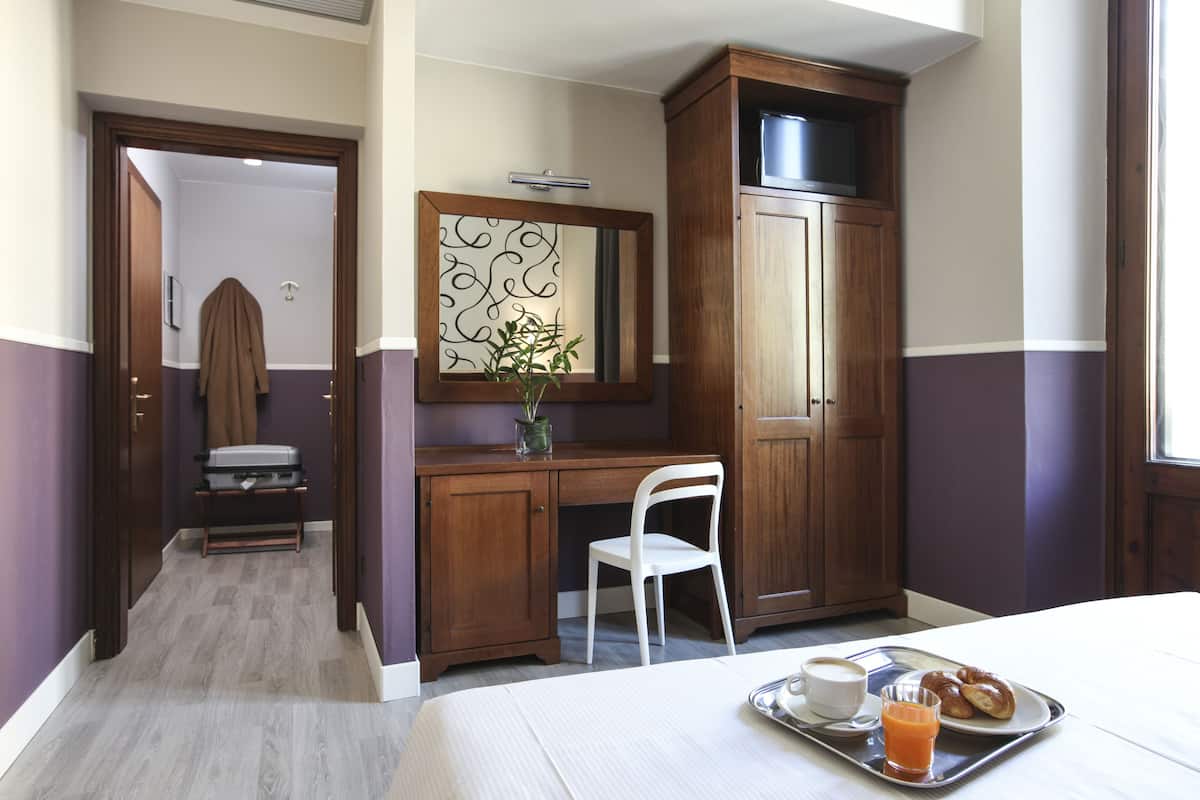 A hotel room with a bed, a desk with a chair, a small table with a tray of breakfast, and a mirror on the wall.