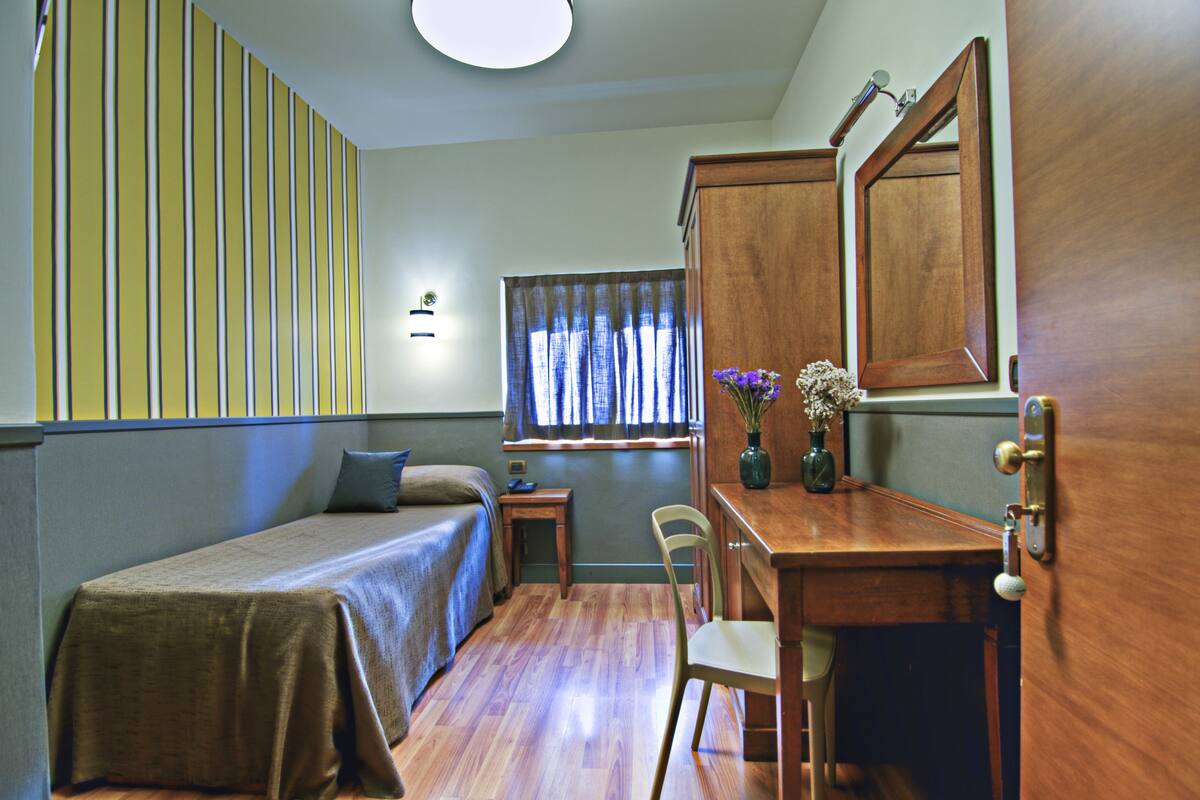 A room with a bed, a desk, a chair, a window with curtains, and a mirror.