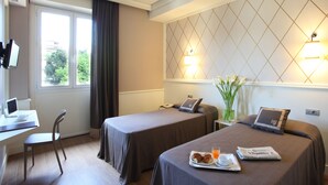 Hotellihuone, jossa on kaksi sänkyä, työpöytä kannettavalla tietokoneella, tuoli, tarjotinalus, jossa on croissantteja ja mehua, sekä sanomalehti.