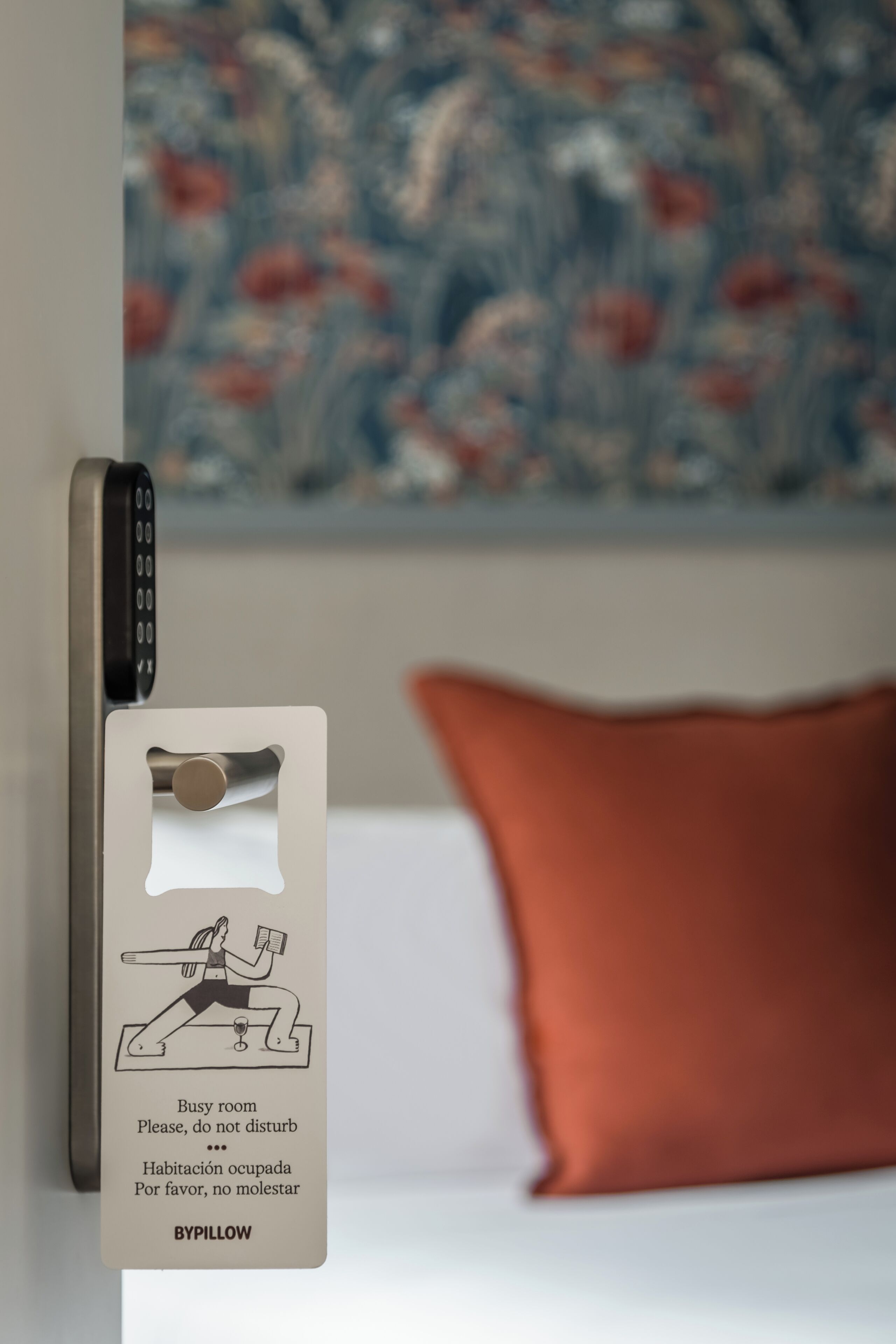 Een hotelkamer met een bed, een rood kussen, een aan de muur bevestigd telefoon en een bord met een yoga-illustratie en tekst in verschillende talen.