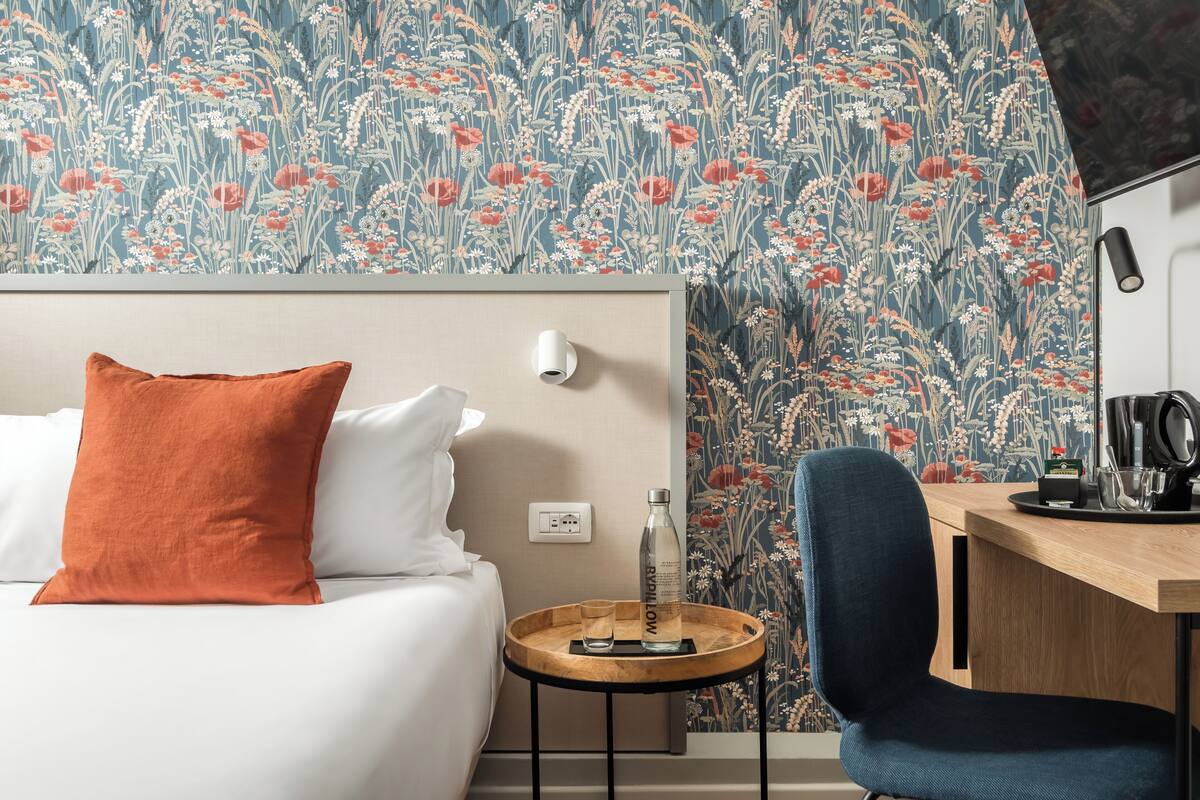 A hotel room with a bed, a desk, a chair, a small round table, a bottle, and a cup.
