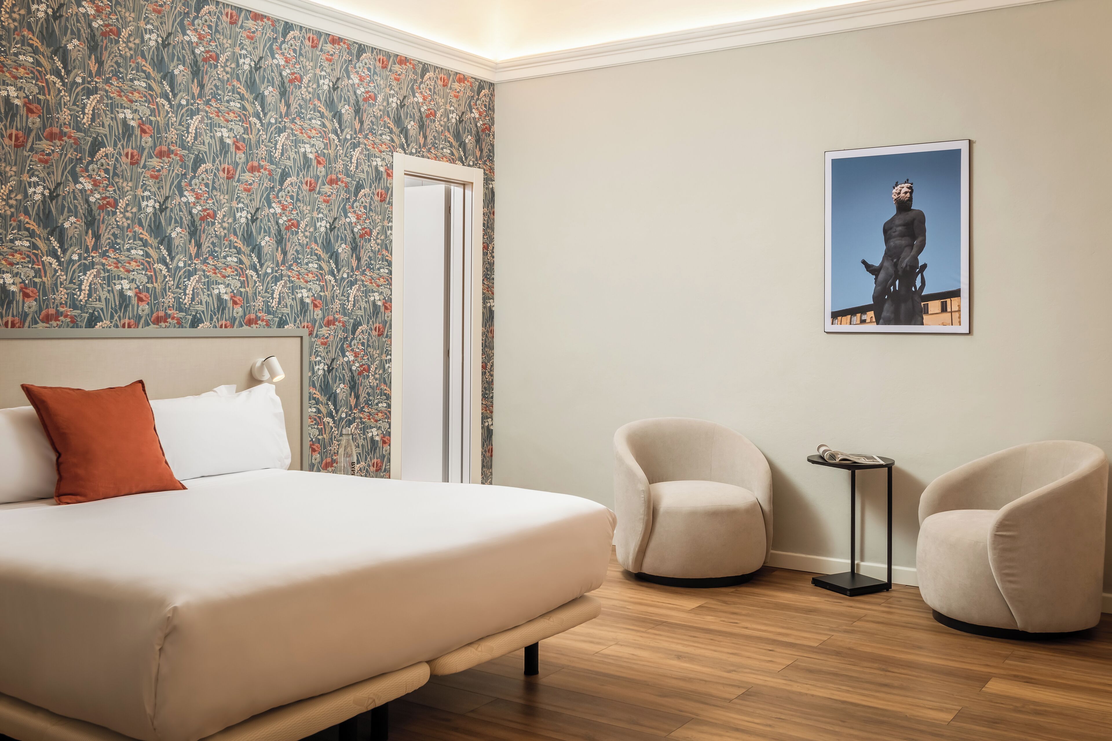 A bedroom with a bed, two armchairs, a small table, a door, and a wall with a floral wallpaper and a framed picture.