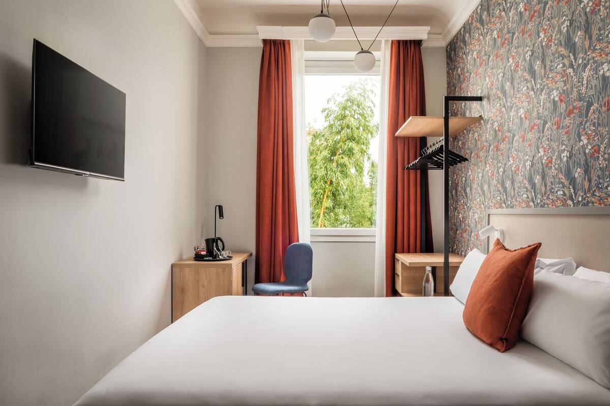 A hotel room with a bed, a desk, a chair, a TV, and a window with curtains.