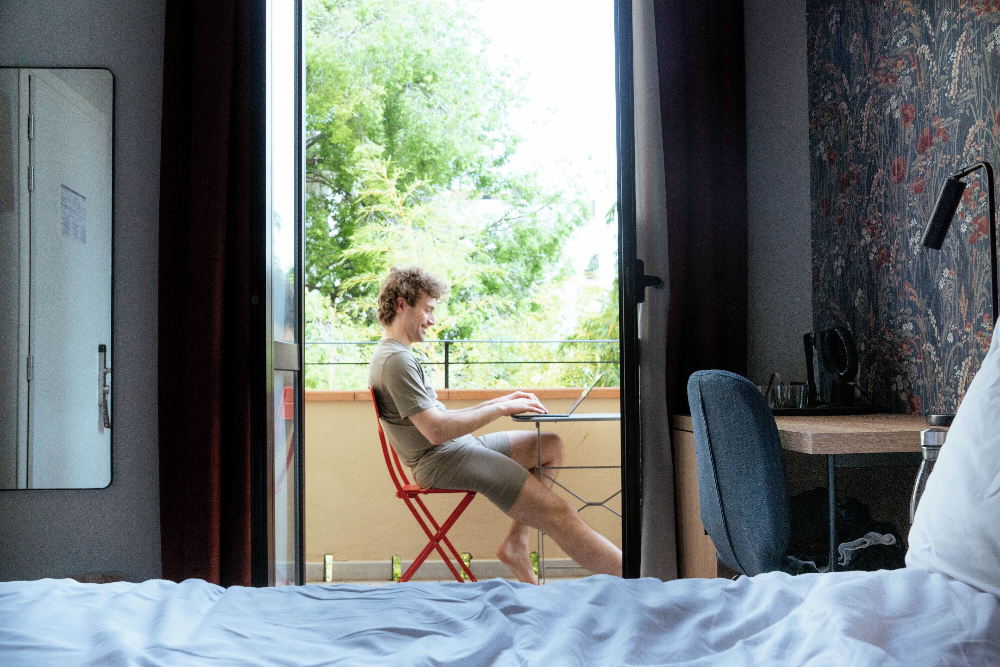 A hotel room with a bed, a desk with a laptop, a chair, and a view of greenery outside.