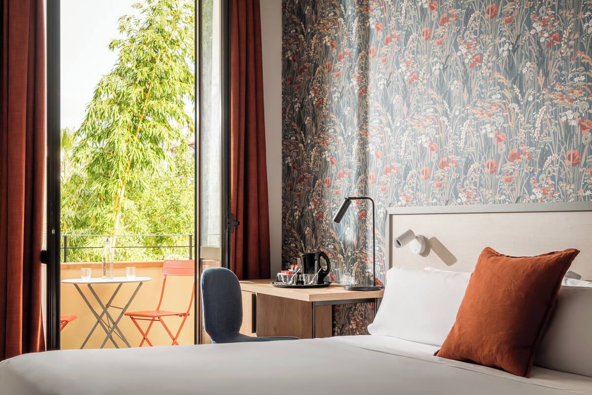 A hotel room with a bed, a desk, a chair, a table, and a view of greenery outside.