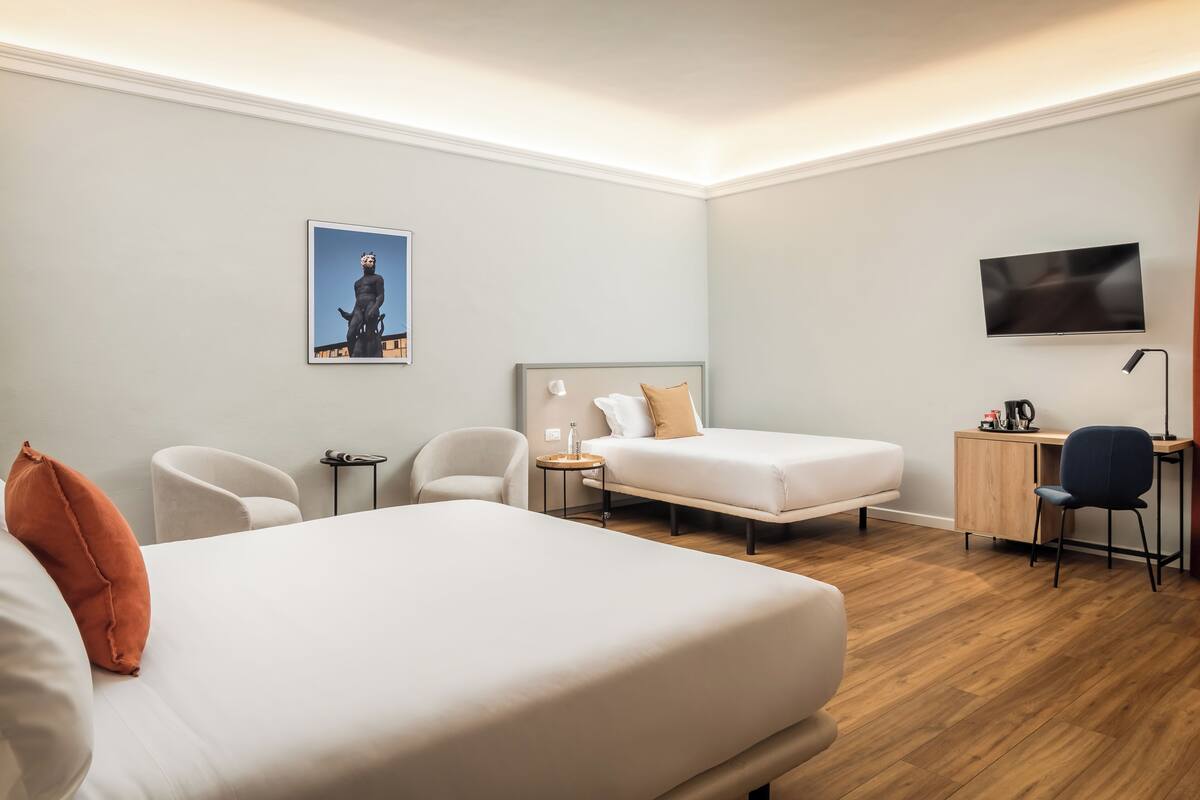 A hotel room with a large bed, a smaller bed, a desk, a chair, a TV, and a statue on the wall.