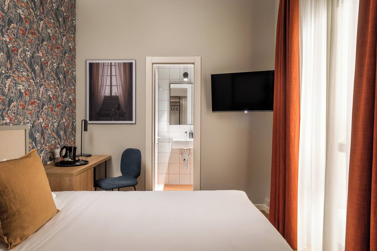 A hotel room with a bed, a desk with a chair, a television, and a bathroom visible through an open door.