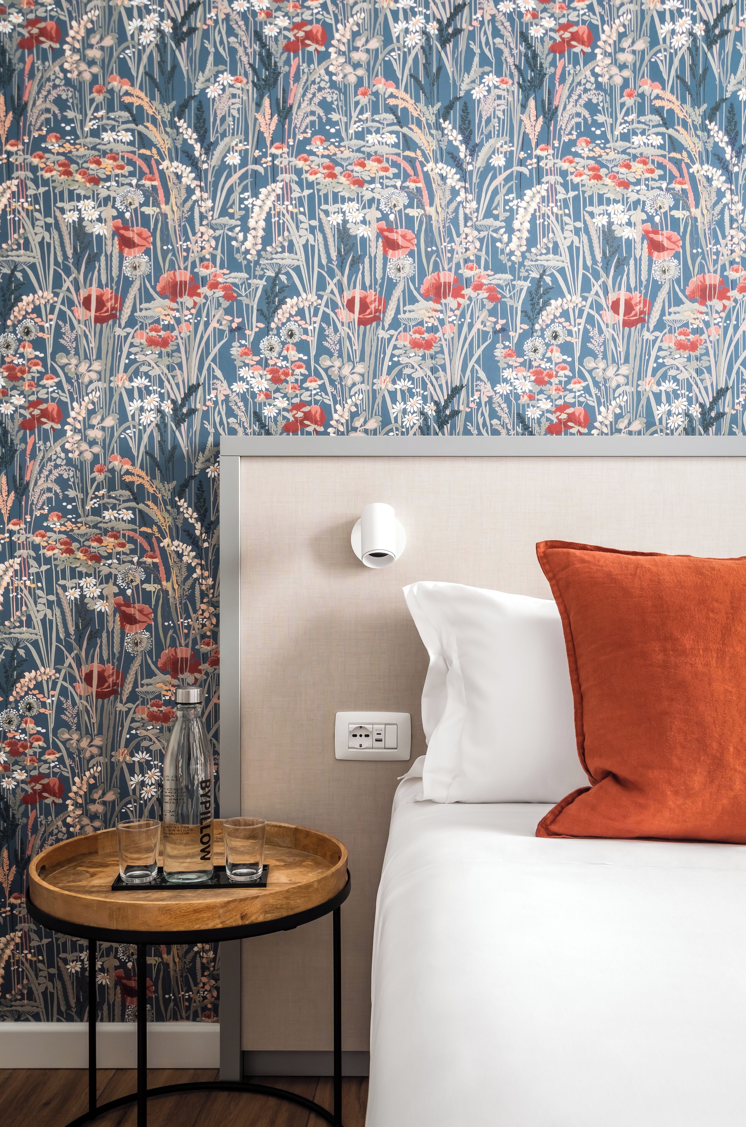 Une chambre d’hôtel avec un papier peint à motifs floraux, une table de chevet avec des verres et une bouteille, un oreiller blanc et un oreiller orange.