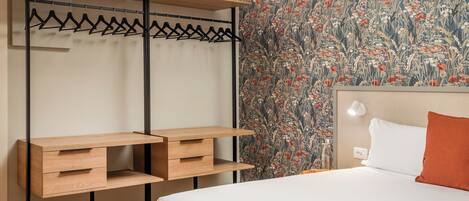 A bedroom with a bed, a wooden desk with drawers, a hanging rack, and a floral wallpaper.