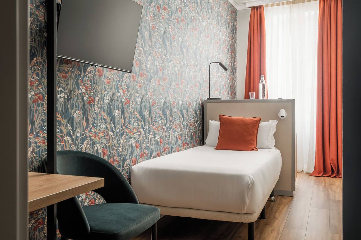 A hotel room with a bed, a desk, a chair, a TV, and a window with curtains.