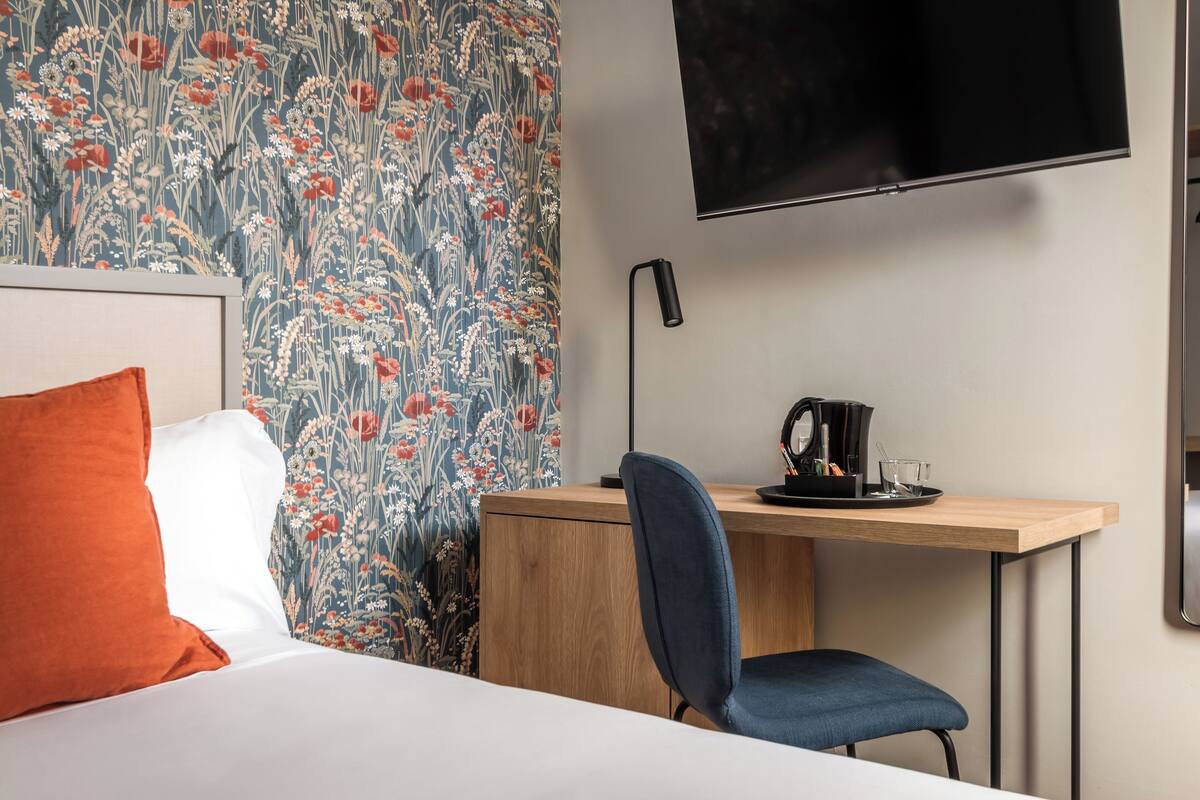A hotel room with a bed, a desk with a chair, a flat-screen TV, and a floral wallpaper.