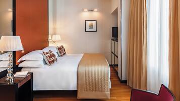 Quarto de hotel com cama, abajures, uma cadeira vermelha, televisão e janela com cortinas.
